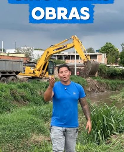 Homem de camisa azul em frente a uma escavadeira que trabalha na margem de um córrego.