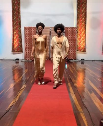 Duas pessoas desfilam em uma passarela com tapete vermelho, vestidas de dourado.